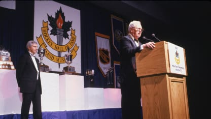 Canadian hockey player and coach Bud Poile (1924 - 2005) speaks at the podium at the Hockey Hall of Fame induction ceremony, September 1990. His son David Poile, general manager of the Nashville Predators, stands behind him. Poile played for several NHL teams, coached for USHL and WHL teams and served as general manager for the Flyers and the Canucks. (Photo by Bruce Bennett Studios via Getty Images Studios/Getty Images)