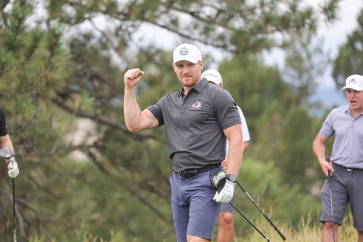 Gabriel Landeskog, wearing a black Colorado Avalanche polo, light blue cap, and golf glove, raises his fist in a celebratory gesture while holding a golf club. He is outdoors on a golf course with trees in the background, surrounded by other players, one of whom is smiling and holding a club.