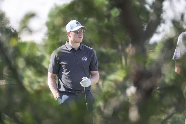 Nathan MacKinnon, wearing a black Colorado Avalanche polo, light blue cap, and sunglasses, stands outdoors holding a golf club. He is framed through blurred greenery, appearing focused, with trees and a bright, sunny sky in the background.