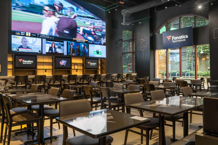 Photo of the inside of the Fanatics Sportsbook. Multiple dining tables and chairs are placed on the floor with bar seating along the back wall. A large screen and many smaller tv screens are lined up on the large back wall.