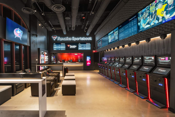 Photo of the inside of the Fanatics Sportsbook. Multiple TV screens and betting kiosks are lined up on the wall to the left. Lounge seating is placed along the wall to the right.