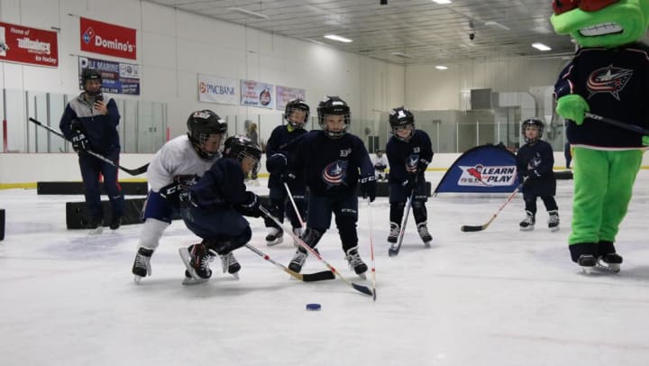 Photo of a group of young children playing hockey alongside Blue Jackets mascot, Stinger, in Blue Jackets hockey gear during a Learn To Play clinic.