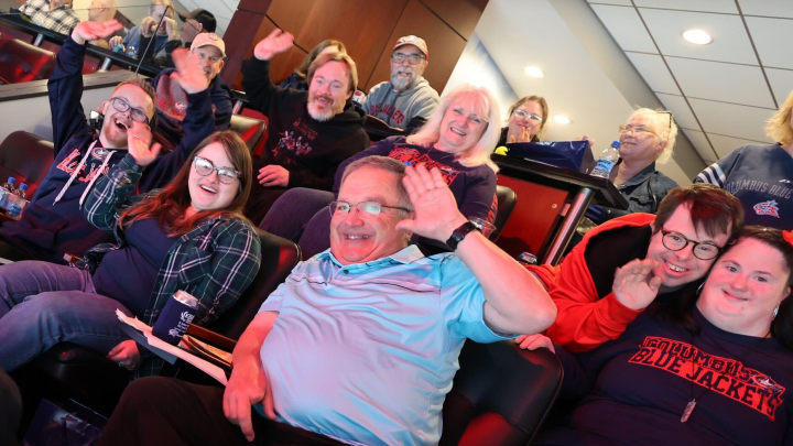 Photo of a group of people waving to the camera while sitting in Zach Werenski's suite at Nationwide Arena during a game.