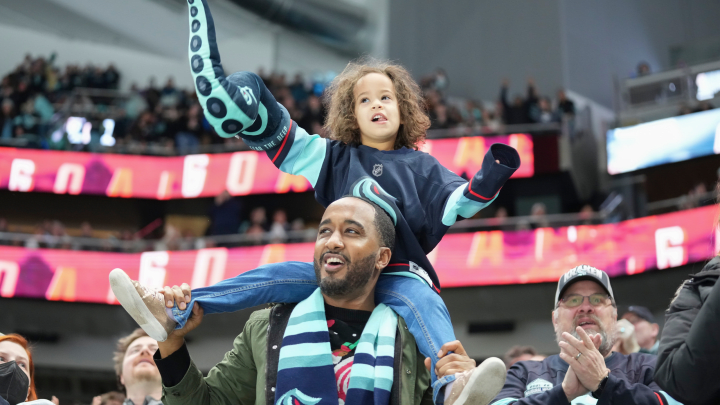 photo of an adult man with a child wearing a dark blue kraken jersey on his shoulders