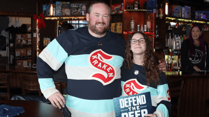 photo of kraken fans wearing kraken jerseys at a bar with hockey on the tv behind them