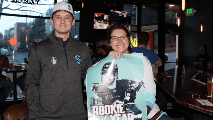 photo of kraken fans wearing kraken gear at a bar with hockey on the tv behind them holding a matty beniers rookie of the year poster